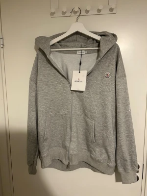 Grå zip hoodie från Moncler - Snygg grå hoodie från Moncler med dragkedja framtill och klassisk Moncler-logga på bröstet. Hoodien har huva, ribbade muddar och två fickor fram. Tillverkad i mjuk bomullsblandning som känns skön mot huden. Perfekt för en avslappnad och trendig look.