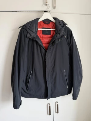 Massimo Dutti marinblå jacka - Snygg jacka från Massimo Dutti med röd innerfoder och huva. Jackan har två dragkedjeförsedda fickor framtill och quiltad baksida. Har en modern, sportig look och är perfekt till våren