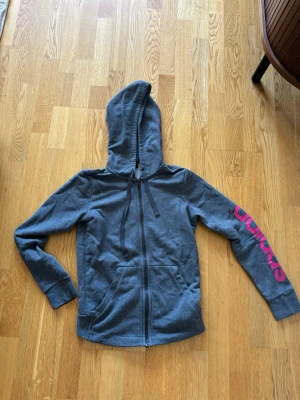 Grå Adidas hoodie med dragkedja - Snygg grå hoodie från Adidas med dragkedja framtill och stor huva. På vänster ärm finns ett rosa Adidas-tryck som ger en sportig vibe. Hoodien har fickor på magen och ribbade muddar vid ärmslut och nederkant. Perfekt för chill eller träning.
