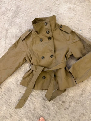 Kort beige trenchcoat med bälte - Stilren kort trenchcoat från Stradivarius i beige bomullsmix. Jackan har dubbelknäppning, axelklaffar och ett matchande midjebälte för snygg passform. Perfekt för lager på lager och trendig look. Klassisk krage och långa ärmar.