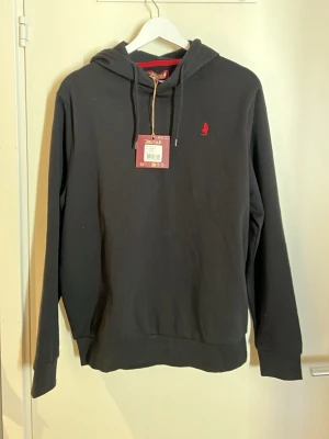 Svart hoodie från U.S. Polo Assn - Svart hoodie från U.S. Polo Assn med röd logga broderad på bröstet. Klassisk modell med huva och dragsnören samt ribbade muddar vid ärmslut och nederkant. Insidan har röda detaljer och materialet känns mjukt och bekvämt, perfekt för en avslappnad stil.