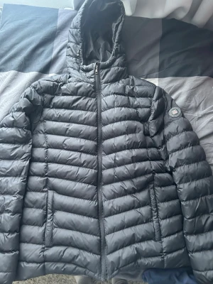 Svart dunjacka med huva från Napapijri - Snygg svart dunjacka från Napapijri med quiltad design och huva. Jackan har dragkedja framtill, två sidofickor och en patch med logga på ärmen. Perfekt för kalla vinterdagar och riktigt skön att bära.