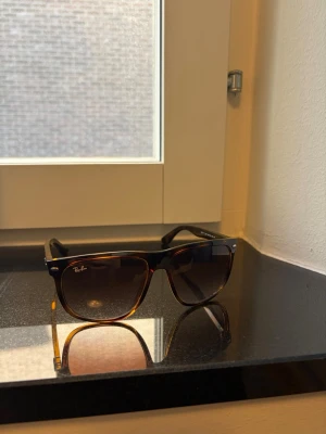 Bruna Ray-Ban solglasögon - Snygga solglasögon från Ray-Ban med klassisk brunsköldpaddsmönstrad båge och mörka glas. Ikonisk logga på sidan och tidlös design som passar till alla outfits. Perfekta för soliga dagar och ger en cool vibe.