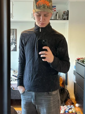 Svart halvzip tröja med emblem - Svart långärmad tröja med halvzip och hög krage. Tröjan har ett diskret emblem på vänster ärm och ribbade muddar vid ärmslut och nederkant. Perfekt för en avslappnad och cool look.