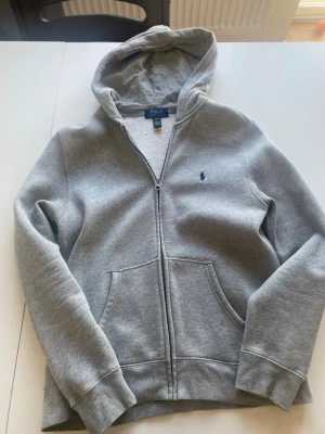 Grå hoodie från Polo Ralph Lauren L(14-16) - Snygg grå hoodie med dragkedja från Polo Ralph Lauren. Klassisk design med blå logga broderad på bröstet. Storlek L(14-16) (junior). Skulle säga att stoleken motsvarar xs/s. Min son köpte den ny på NK förra året. Den är i ett fint skick. 
