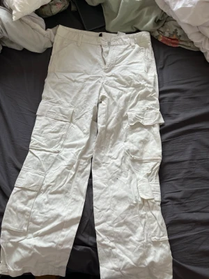 Vita cargobyxor från H&M Divided - Snygga vita cargobyxor från H&M Divided med flera stora fickor på sidorna och raka ben. Byxorna har bälteshällor och stängs med knapp och dragkedja. Perfekta för en avslappnad och trendig stil.