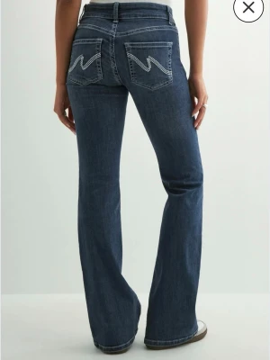 Mörkblå low waist bootcut jeans från Nelly  - Säljer ett par mörkblå low waist bootcut jeans med vita kontrastsömmar med zigzag detalj på bakfickorna från Nelly. Har inte använt byxorna och har även prislappen fortfarande på. 