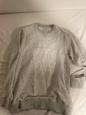 Grå sweatshirt från Les Deux - Snygg ljusgrå sweatshirt från Les Deux med broderad text 'Palais Royal' på bröstet. Tröjan har rund halsringning, ribbade muddar vid ärmslut och nederkant samt en avslappnad passform. Perfekt för en casual look. Väldigt bra skick. Inga fläckar och inte utslitet på något sätt. 