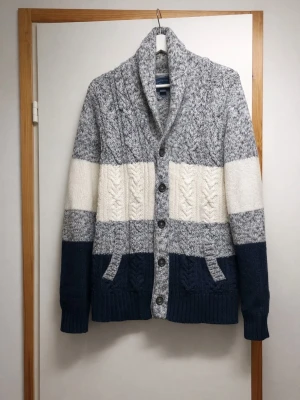 Stickad Cardigan blått, grått och vitt - Säljer en chunky stickad kofta med breda blockränder i marinblått, gråmelerat och vitt. Koftan har grov kabelstickning, stora knappar framtill, två fickor och en hög krage. Perfekt för kalla dagar och riktigt mysig att ha på sig.