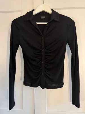 Svart långärmad topp/skjorta med rynk - Trendig svart långärmad topp/skjorta med krage och knappar framtill. Toppen/skjortan har snygg rynkdetalj längs knappraden som ger en cool look. Tillverkad i ett mjukt och stretchigt material som sitter snyggt på kroppen. Perfekt att styla med jeans eller kjol.