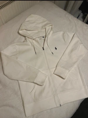 Vit hoodie från Ralph Lauren - Säljer en stilren vit hoodie från Ralph Lauren med dragkedja framtill och klassisk huva med snören. Tröjan har två fickor och den ikoniska lilla hästloggan broderad på bröstet. Perfekt för en clean och avslappnad look.