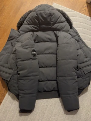 Grå dunjacka från Supply med pälshuva - Snygg grå dunjacka från Supply med stor huva och fuskpälsdetalj. Jackan har ett stilrent badge på ärmen och breda kanaler för extra värme. Perfekt för kalla vinterdagar och har en modern, boxig passform.