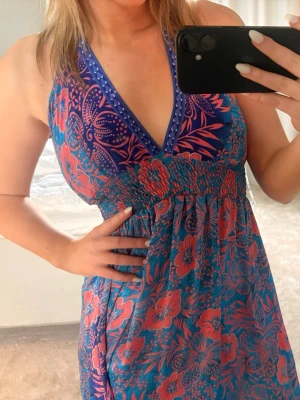 Blommig långklänning med öppen rygg - Snygg långklänning med blå och rosa blommigt mönster som jag köpte på en marknad i cypern! djup v-ringning och öppen rygg med knytband. Klänningen har smock i midjan och tunna axelband. Perfekt för sommarens varma dagar.