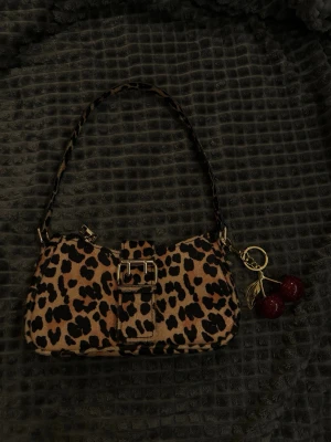 Leopardmönstrad axelväska med körsbär - Cool handväska i leopardmönstrat tyg med guldfärgat spänne och matchande axelrem. Väskan har en dekorativ körsbärsnyckelring i rött och guld. Perfekt för dig som vill sticka ut med ett djurmönster och snygga detaljer.