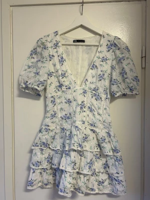Blommig kortklänning från Zara - Superfin kortklänning från Zara med blått blommönster på vit botten. Klänningen har v-ringning, puffärmar och volangdetaljer i kjolen. Perfekt för sommarens alla tillfällen!