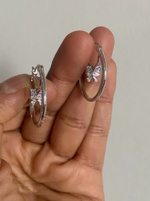s925 sterling Silvriga hoops med fjärilar - Snygga silvriga örhängen i hoop-modell med hängande fjärilar dekorerade med små glittrande stenar. Fjärilarna har en lätt lila ton i stenarna som ger extra bling. Perfekta för dig som gillar trendiga och söta accessoarer. sterling silver pläterat 