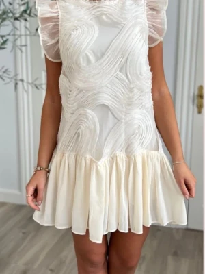 Vit kortklänning med volang och struktur - Superfin vit kortklänning med volangkant nertill och transparenta, korta ärmar. Klänningen har ett unikt strukturerat mönster och pärldetaljer vid halsringningen. Perfekt för studenten eller bara sommarens tillfällen och ger en romantisk känsla. Färgen är mer cream vit.