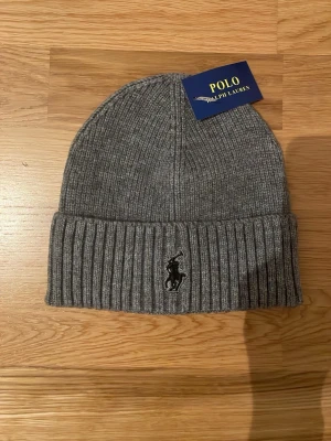 Grå ribbstickad mössa Polo Ralph Lauren - Snygg grå ribbstickad mössa med Polo Ralph Lauren logga. Perfekt accessoar för kyliga dagar och enkel att matcha med olika outfits.