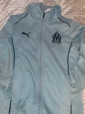 Ljusblå Marseille hoodie från Puma - Ljusblå hoodie med dragkedja från Puma, officiell Marseille-tröja med klubbens logga och texten 'DROIT AU BUT' på bröstet. Mörkblå detaljer längs sidorna och MARSEILLE tryckt på ryggen. Perfekt för fotbollsfans och träning.