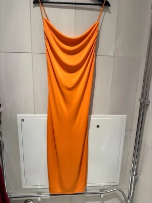 Orange långklänning med smala axelband - Snygg orange långklänning med smala axelband och draperad urringning. Klänningen har en rak passform och är tillverkad i ett mjukt, slätt material som blir figursytt efter din kropp. Perfekt för dig som vill sticka ut med färg.