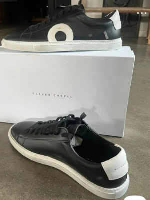 Svarta sneakers från Oliver Cabell - Ett par helt oanvända Svarta sneakers i läder från Oliver Cabell med vit sula, vit hälkappa och en stor vit cirkeldetalj på sidan. Klassisk låg modell med svarta skosnören och minimalistisk design. Perfekta för dig som gillar stilrena och trendiga skor. Ny pris runt 3000kr då tull var dyr, de kommer med box och dustbag 