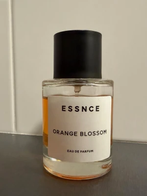ESSNCE Orange Blossom parfym - Fräsch Eau de Parfum från ESSNCE med doften Orange Blossom. Flaskan är cylindrisk i genomskinligt glas med svart matt kork och vit etikett med svart text. Innehållet har en varm orange färg som ger en lyxig känsla.