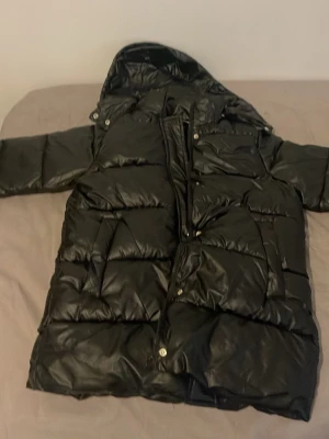 Svart lång dunjacka från Moncler - Säljer en svart lång dunjacka från Moncler med huva och stora, quiltade partier. Jackan har dragkedja och tryckknappar framtill samt en bröstficka med lock. Perfekt för kalla vinterdagar och riktigt snygg streetstil.