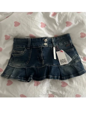 Mörkblå jeanskjol med volang - Mörkblå jeanskjol från Urban Outfiters BDG med volangkant och dubbla knappar framtill. Kjolen har klassiska fickor fram och bak, där bakfickorna har dekorativa sömmar som formar hjärtan. Perfekt för en trendig och avslappnad look. Återpublicerad annons.