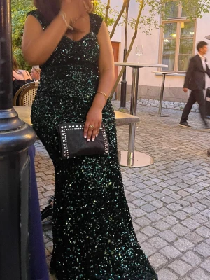 Balklänning  - En fantastisk långklänning i mörkgrönt med glittriga paljetter över hela klänningen. Klänningen har breda axelband och figurnära passform som framhäver siluetten. Perfekt för bal eller festliga tillfällen.