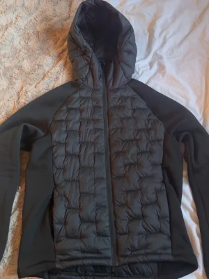 Svart pufferjacka med huva - Snygg svart pufferjacka från Everest, Stadium, med quiltad framsida och rygg samt släta ärmar. Jackan har hel dragkedja och en stor huva för extra värme. Perfekt för kalla dagar och har en sportig look. Materialet är syntet och jackan är lätt vadderad. Använd ca 2-3 gånger, sedan har den bara hängt i garderoben. Den är i nyskick och har i princip inga tecken på användning. Ny pris är 799.