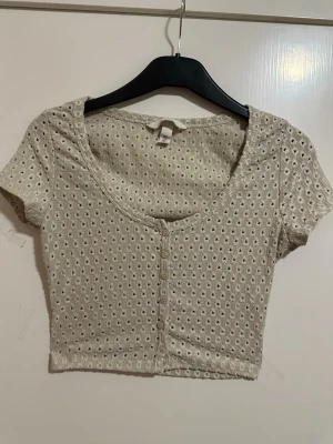 Beige croppad topp med knappar strl: S - Söt beige croppad topp med korta ärmar och dekorativa knappar framtill. Toppen har ett hålmönster i tyget och en djup rundad ringning. Perfekt för dig som gillar en enkel men ändå detaljrik stil.