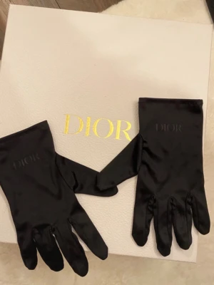 Svarta satin-handskar från Dior - Eleganta svarta handskar från Dior i mjukt satinmaterial. Handskarna har en diskret Dior-logga på ovansidan och en klassisk, slät design som ger en lyxig känsla. Perfekta för dig som vill addera en exklusiv touch till din outfit. Fått i present men aldrig använda. Obs, stora lådan ingår ej, endast handskarna!