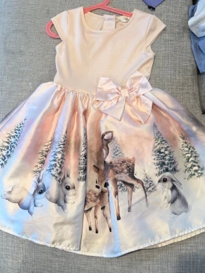 Söt ljus klänning med djurmotiv - Ljus klänning med rund hals och korta ärmar, dekorerad med ett stort sidenliknande rosett i midjan. Kjolen har ett sagolikt tryck med rådjur, kaniner och granar i mjuka pastellfärger. Perfekt för speciella tillfällen.