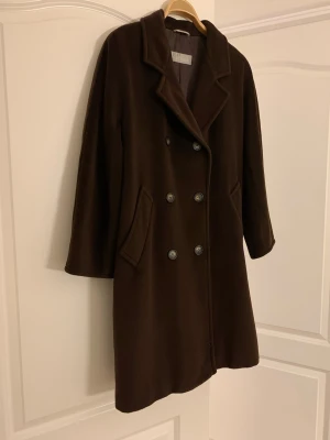 Mörkbrun vintage kappa från MaxMara - Stilren mörkbrun kappa från MaxMara i ullmix med dubbelknäppning och klassisk krage. Kappan har två snedställda fickor och en rak, något längre passform. Perfekt för dig som vill ha en tidlös och elegant look.