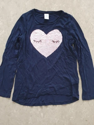 Tröja - Mörkblå långärmad tröja med stort rosa hjärta i paljetter på bröstet. Hjärtat har ett sött motiv med stängda ögon. Perfekt för dig som gillar detaljer och vill sticka ut lite extra. Mjuk och skön känsla, troligtvis i bomull eller polyester.