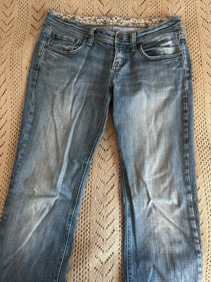 Blå bootcut jeans med slitningar - Snygga blå bootcut jeans med ljusa slitningar framtill och klassisk femficksdesign. Jeansen har normal midja och dragkedja med knapp. Perfekta för en avslappnad och trendig look.