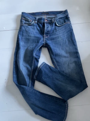 Blå jeans från Nudie Jeans - Säljer ett par klassiska blå jeans från Nudie Jeans med orange sömmar och broderade bakfickor. Jeansen har rak passform och är tillverkade i denim av bomull. Perfekta för en avslappnad och stilren look.