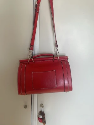 Röd handväska med axelrem - Snygg röd handväska i läderimitation med justerbar axelrem och handtag. Väskan har vita kontrastsömmar, dekorativa spännen och ficka både fram och bak. Perfekt för dig som vill ha en stilren och trendig accessoar.