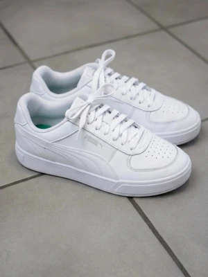 Vita Puma sneakers i läder - Fräscha vita sneakers från Puma i klassisk low-modell. Skorna är tillverkade i slätt läder med perforerade detaljer på tån och har vita skosnören. Diskret Puma-logga på sidan och bekväm innersula. Perfekta för en clean och stilren look.