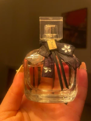 Yves Saint Laurent Mon Paris parfym 50ml - Elegant parfymflaska från Yves Saint Laurent, Mon Paris. Flaskan är genomskinlig med en fyrkantig form och dekorerad med en svart rosett med små vita blommor och ett guldfärgat YSL-emblem. Innehåller 50 ml Eau de Parfum.
