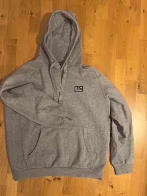 Grå hoodie från EA7 Emporio Armani - Snygg grå hoodie från EA7 Emporio Armani med klassisk huva och dragsnören. Mjuk bomullsblandning och känguruficka framtill. Diskret EA7-logga på bröstet ger en clean och stilren look. Perfekt för dig som gillar streetwear och enkel design.  Pyttelitethål 