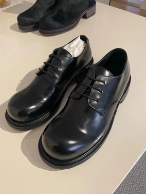 Derby skor Acne Studios - Stilrena svarta loafers i blankt läder från Acne Studios. Klassisk rund tå, låg klack och robust sula i trä med gummidetalj. Perfekta för en clean och dressad look. Skorna har minimalistisk design och svarta snören.