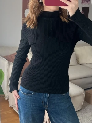 Svart ribbstickad tröja med hög krage - En svart ribbstickad tröja med lång ärm och hög krage. Tröjan har en figurnära passform och är gjord i ett mjukt stickat material som passar perfekt till jeans. Enkel och stilren design utan synliga loggor eller mönster.