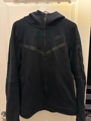 Svart Nike hoodie med dragkedja - Säljer en svart hoodie från Nike med hel dragkedja och huva. Hoodien har diskret Nike-logga på bröstet, snygga detaljer i form av svarta linjer över bröstet och en praktisk ficka med dragkedja på ärmen. Endast använd fåtal gånger.