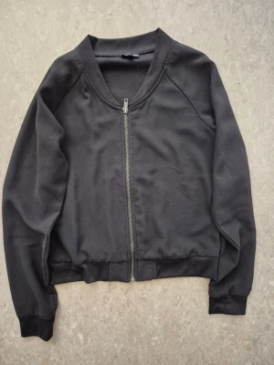 Bomberjacka - Stilren svart bomberjacka med rak passform och silverfärgad dragkedja framtill. Jackan har ribbade muddar vid ärmslut och midja samt rundad krage. Perfekt för en enkel och clean look. Har en fläck.