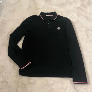 Moncler tröja  - Säljer en stilren svart långärmad pikétröja från Moncler. Den har en klassisk krage med röd och vit rand samt knappar framtill som symboliserar moncler färger. Perfekt för både vardag och lite finare tillfällen. Moncler-loggan är broderad på bröstet. Passar perfekt till höst och vår! Ny pris 2999kr vårt pris 999. 