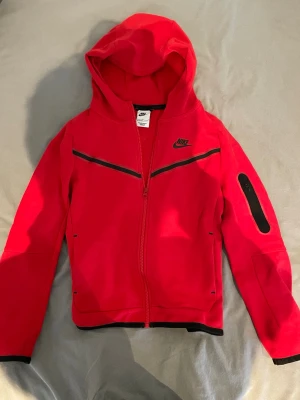 Äkta Röd Nike tech med dragkedja - Snygg röd Nike tech med hel dragkedja och svarta detaljer. Huva och ficka på ärmen med dragkedja ger en sportig vibe. Tillverkad i mjukt material, perfekt för chill eller träning. Nike-logga på bröstet och coola svarta linjer framtill. Storlek 147-158. Mycket fint skick på tröjan. Nypris ca 800 kr.