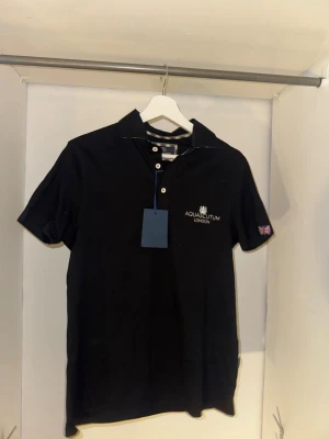 Svart pikétröja från Aquascutum - Klassisk svart pikétröja från Aquascutum London med korta ärmar och krage. Tröjan har broderad logga på bröstet och en liten brittisk flagga på ärmen. Stängs med knappar framtill och är tillverkad i mjuk bomull.