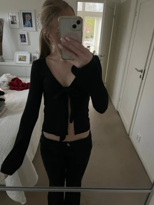Svart croppad topp med knyt - Säljer en svart croppad topp med lång ärm och v-ringning. Toppen har ett snyggt knyt framtill som ger en cool look. Perfekt för dig som gillar trendiga och stilrena plagg.