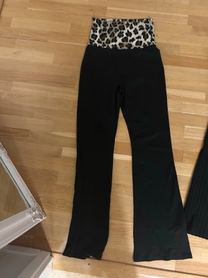 Svarta leggings med leopardmönstrad midja - Coola svarta yoga pants i leopardmönster. Modellen har utsvängda ben och bred linning som ger en snygg siluett. Perfekta för dig som vill sticka ut med en trendig detalj. Materialet är stretchigt och bekvämt.